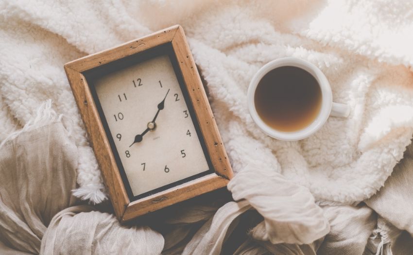 a clock beside a cup of coffee on a bed