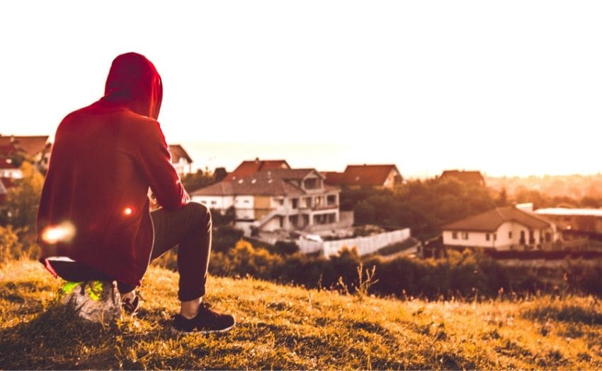 person wearing a red hoodie sitting on a grassfield overlooking some houses