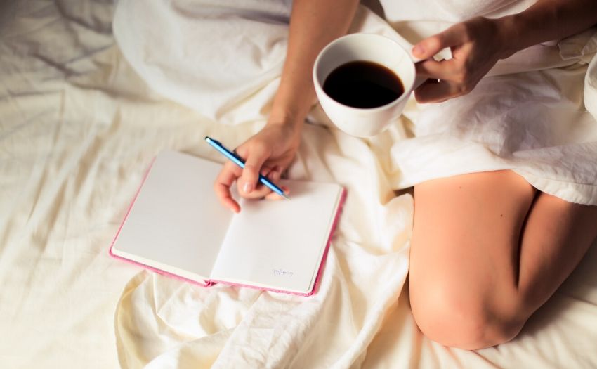 How to start journaling feature image - woman sitting on bed with a notebook and a cup of coffee