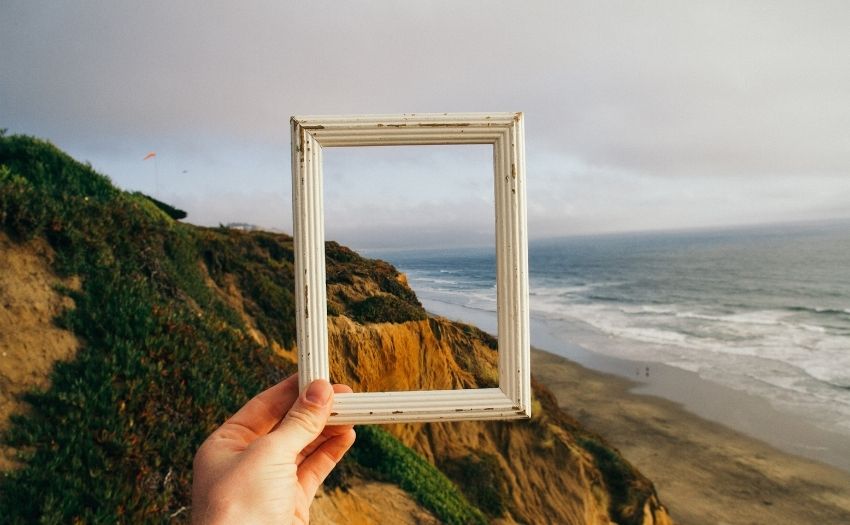 a hand holding up a white wooden picture frame in front of landscape