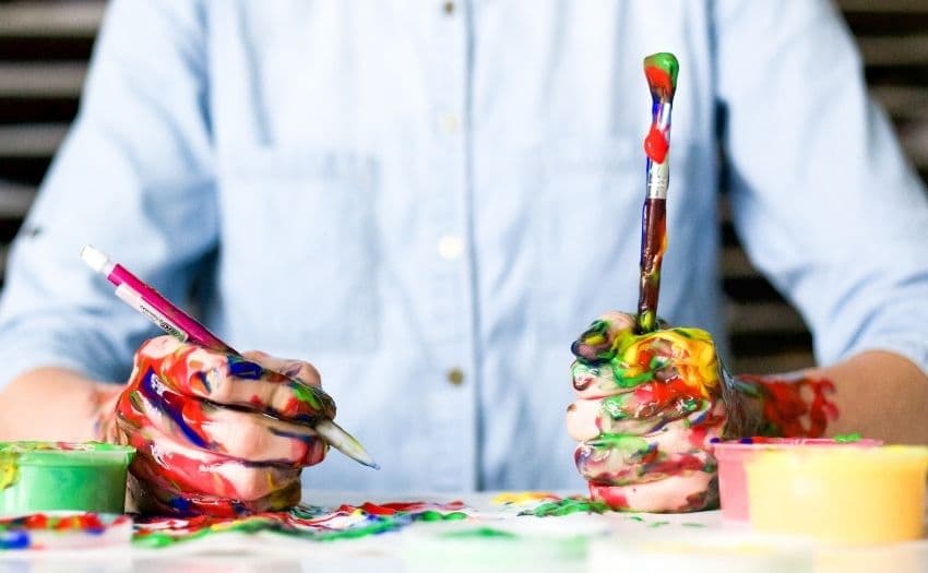 person holding paintbrushes and has paint all over their hands